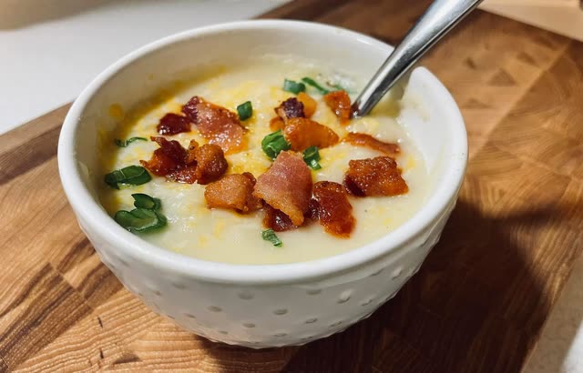 A white bowl of creamy soup topped with crispy bacon, cheddar and green onion on a wooden board
