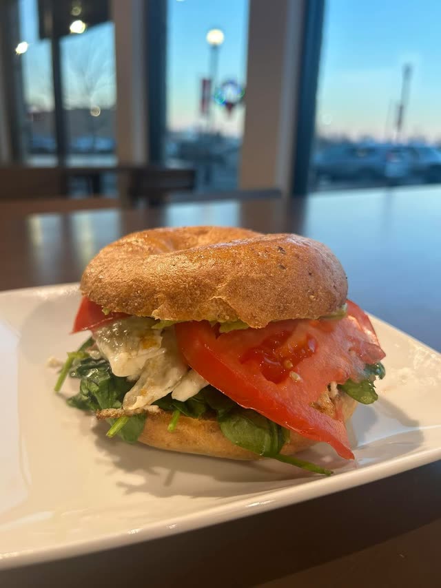 A bagel sandwich with spinach, egg and tomato on a white plate by the café window