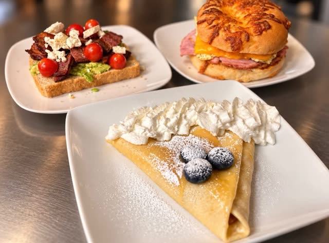 Three plates on a steel counter: avocado toast topped with bacon, cherry tomatoes and crumbled feta; a ham and cheese bagel sandwich; and a folded crepe with whipped cream, powdered sugar and blueberries