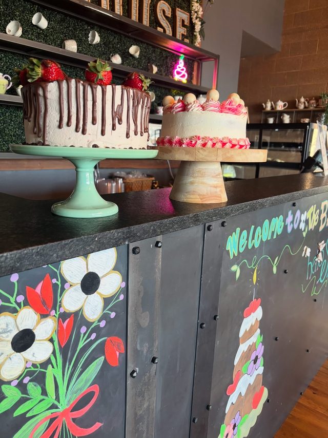 Two cakes on stands on the counter, a chocolate-drip cake topped with strawberries and a white cake piped with pink swirls, above the hand-painted counter front with flowers and a layered cake