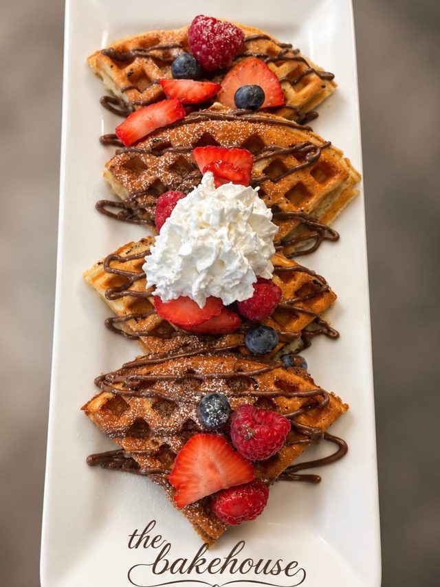 Waffle wedges on a long white plate printed with “the bakehouse”, with chocolate drizzle, strawberries, raspberries, blueberries and whipped cream