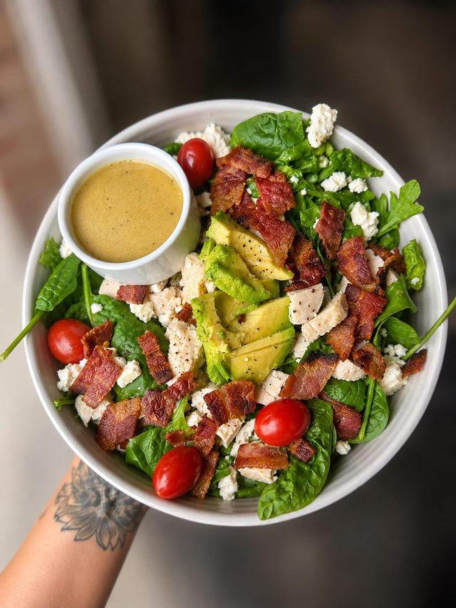 A hand holding a bowl of spinach salad with crispy bacon, avocado, cherry tomatoes, feta, chicken and a cup of dressing